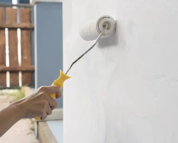 Mano aplica capa de imprimación blanca a una pared con un rodillo de pintura amarillo.