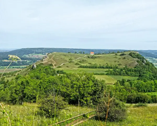 Grüne Hügel und Felder laden zum Walberla wandern ein. Ein kleiner Weg schlängelt sich zum Gipfel mit einem roten Haus.