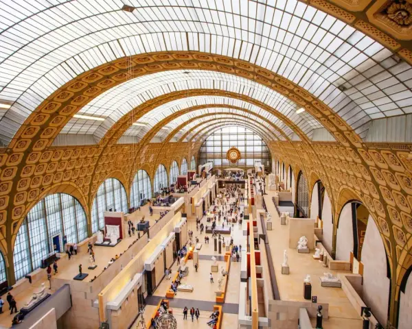 Interior del museo D'Orsay, con su impresionante techo abovedado de cristal y arcos dorados. Gente pasea entre esculturas y exposiciones.