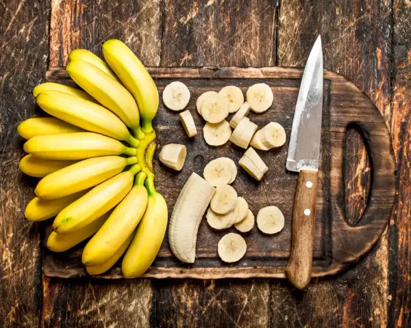 Pęczek żółtych bananów i pokrojone plasterki na desce. Dowiedz się, jak przechowywać banany, by dłużej były świeże.