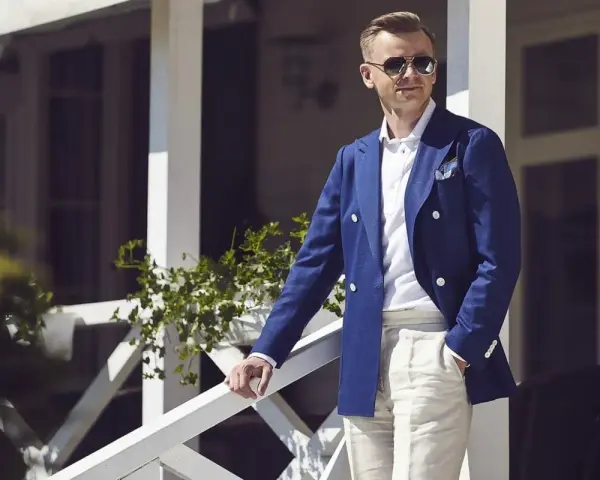Elegancki mężczyzna w granatowej marynarce i beżowych spodniach, w stylu ivy league, stoi na werandzie.