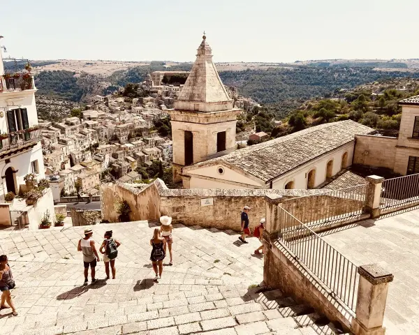 Panoramica di una città barocca in Sicilia orientale, cosa vedere: un borgo arroccato con chiese e case in pietra, turisti che esplorano le scalinate.