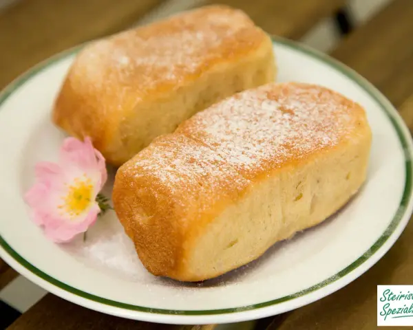Zwei goldbraune Buchteln, bestäubt mit Puderzucker, auf einem Teller mit einer zarten rosa Blüte. Omas Rezept für Buchteln, ein Genuss!