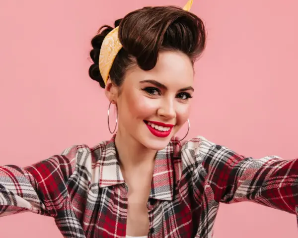 Uśmiechnięta kobieta w stylu pin-up, z burzą brązowych włosów spiętych w kok i żółtą bandaną, w kratę koszuli, robi sobie selfie.