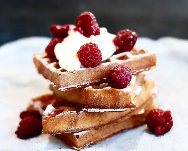 En stapel gyllene belgiska våfflor toppade med vispgrädde och färska hallon, pudrade med florsocker. En härlig frukost eller efterrätt.