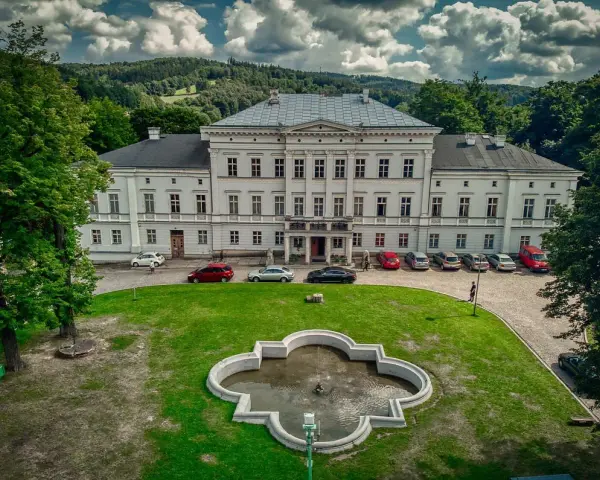 Pałac Jedlinka, biała, okazała budowla z szarym dachem, otoczona zielenią i górami. Przed pałacem fontanna i parking z samochodami.