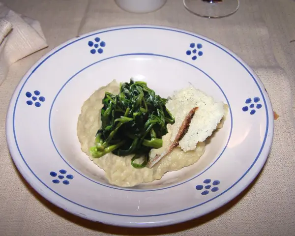 Piatto con fave e cicoria saltata, guarnito con una scaglia di formaggio e un'acciuga.