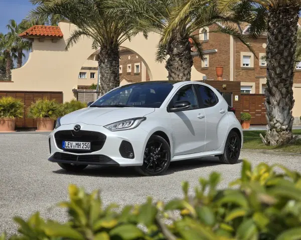 Biała Mazda 2 Hybrid zaparkowana przed budynkiem z palmami. Stylowy miejski samochód gotowy na podróż.