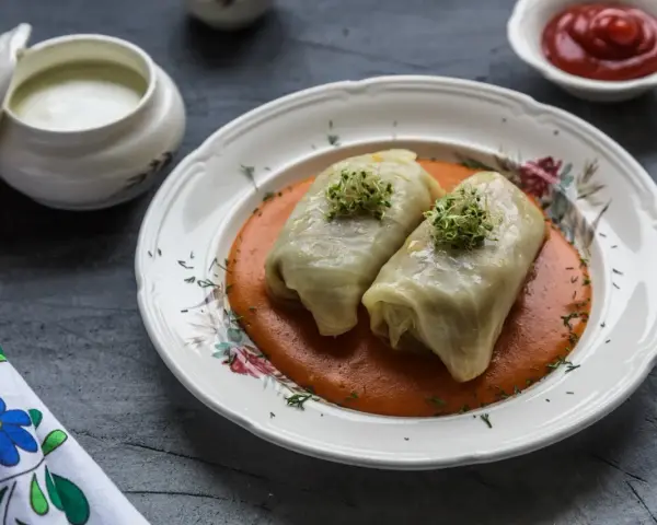 Dwa gołąbki w sosie pomidorowym, udekorowane kiełkami. Pytanie brzmi: czy gołąbki można mrozić? Obok sos śmietanowy i ketchup.