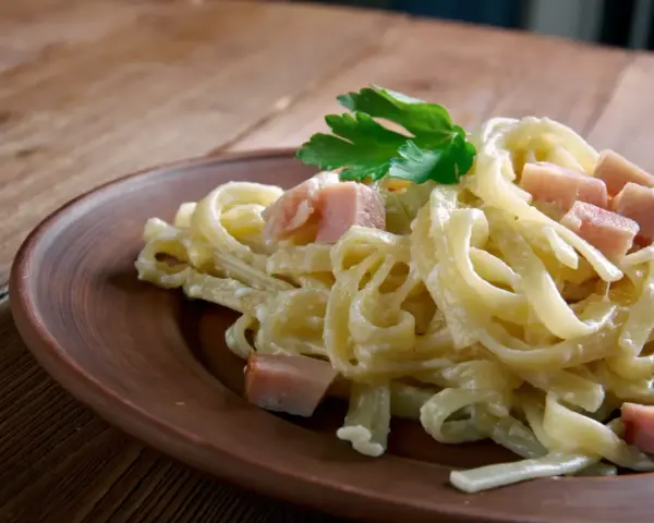 Fettuccine co to? Pyszne, kremowe fettuccine z kawałkami szynki i natką pietruszki na brązowym talerzu.