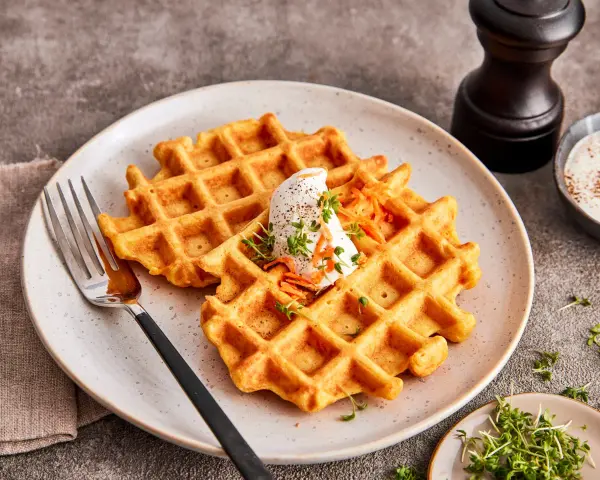 Zwei goldbraune Karotten Waffeln mit einem Klecks Sahne und geraspelten Karotten, garniert mit Kresse.