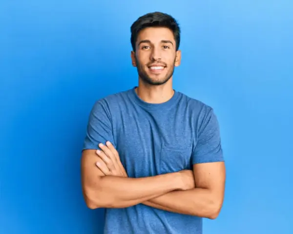 Hombre sonriente con estilos barba poco poblada, brazos cruzados, sobre fondo azul.