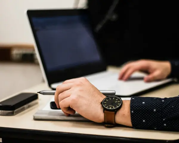 Dłoń z zegarkiem trzyma długopis nad dokumentem, obok laptop. Czas na skargę na przewlekłość postępowania opłatę.