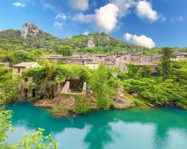 Un borgo incantevole in Umbria, con case in pietra ricoperte di vegetazione che si affacciano su un fiume turchese. Un luogo perfetto per i cammini.