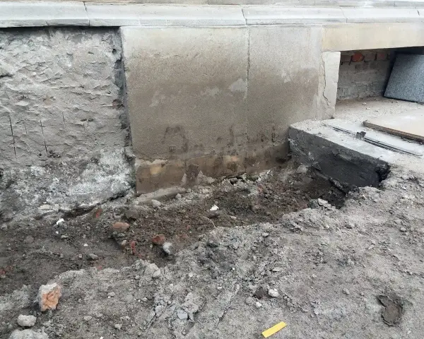 Widoczne ślady osiadania budynku i pęknięcia na cokole. Uszkodzony fragment fundamentu, ziemia i gruz wokół.