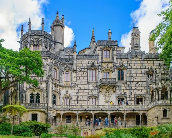 The Quinta da Regaleira estate in Sintra, Portugal, features a stunning palace with intricate stonework and ornate balconies, surrounded by lush greenery under a blue sky.