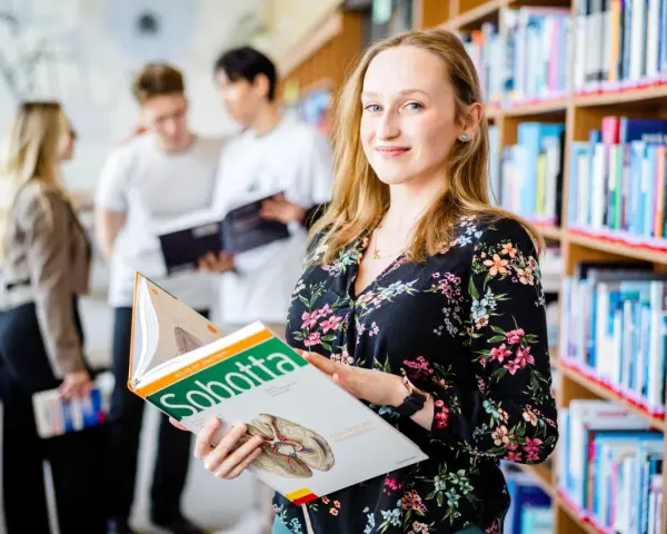 Studentka na Uniwersytecie Wrocławskim, kierunek prawo, przegląda atlas anatomii. W tle inni studenci i regały z książkami.