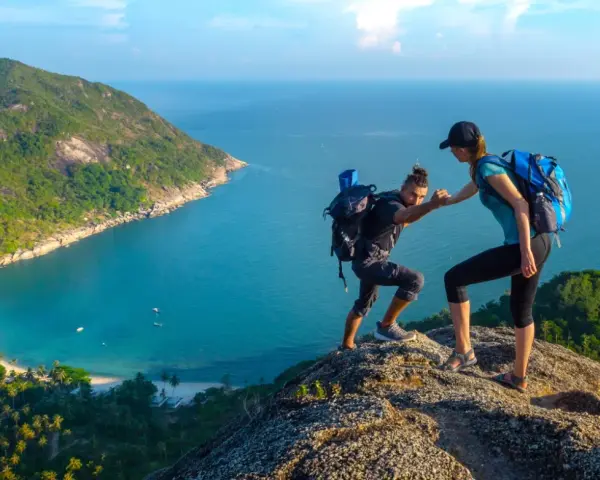 Due escursionisti si aiutano a salire su una scogliera con vista su una baia tropicale. Questo è il trekking cos'è: avventura e collaborazione.