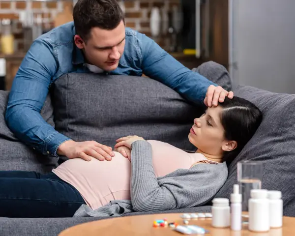 Zmartwiony partner troskliwie dotyka czoła ciężarnej kobiety, która leży na kanapie. Na stoliku leki, sugerujące problemy zdrowotne, być może angina w ciąży.