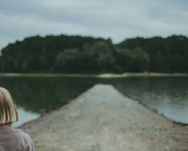 Dziewczyna z blond włosami patrzy na spokojną taflę jeziora, czując uczucie pustki.