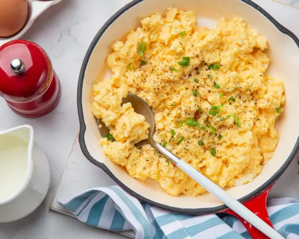 Un plat de oeufs brouillés, parsemé de ciboule et de poivre, servi dans une poêle rouge. Un moulin à poivre rouge et une cruche de lait sont à proximité.