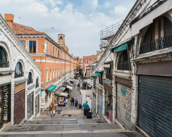 Scendendo le scale del ponte di Rialto a Venezia, si intravede un mercato vivace con negozi e bancarelle.