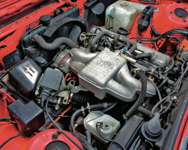 Silniki BMW E30 w czerwonej komorze silnika. Widoczny kolektor ssący z logo BMW, chłodnica, akumulator i inne elementy układu napędowego.
