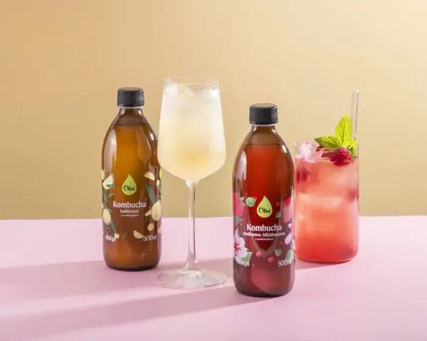 Kombucha co to? Dwie butelki kombuchy Olini: imbirowej i malinowo-hibiskusowej, obok orzeźwiających napojów z lodem.