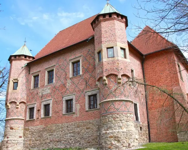 Zamek w Dębnie, ceglana twierdza z kamienną podstawą i zielonymi wieżyczkami, otoczony drzewami pod błękitnym niebem.