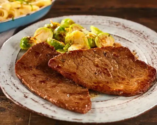 Due fette di paillard di vitello succulento servite con cavolini di Bruxelles arrostiti e un contorno di pasta in una padella.