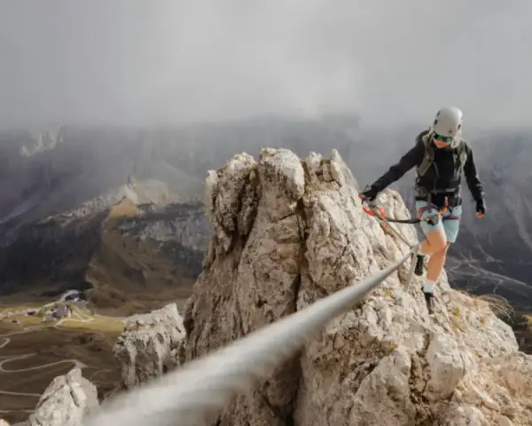 Kobieta w kasku i uprzęży pokonuje via ferrata, balansując na linie nad skalistym zboczem. W tle rozciąga się malownicza dolina.