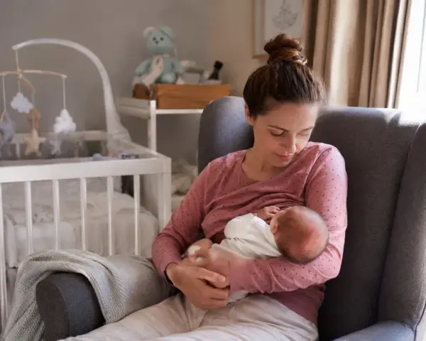 Matka karmi piersią noworodka w fotelu. W tle widać łóżeczko dziecięce. To piękny obrazek z okresu po porodzie.