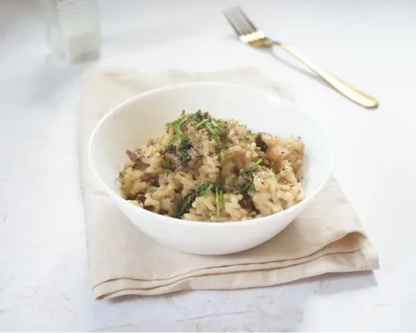 Miseczka kremowego risotto z grzybami, posypana ziołami i pieprzem, obok złoty widelec.