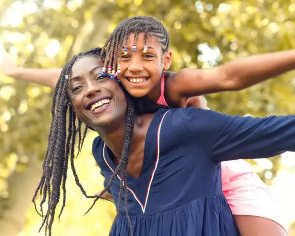 Une femme porte une enfant sur son dos, toutes deux souriantes, avec des arbres en arrière-plan.