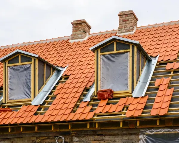 Trwa wymiana okna dachowego. Dach pokryty czerwona dachówką, widoczne drewniane elementy konstrukcji i nowe okna z folią.