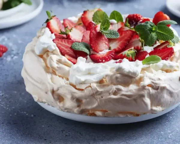 Pyszny tort bezowy z mascarpone i bitą śmietaną, ozdobiony świeżymi truskawkami i miętą.