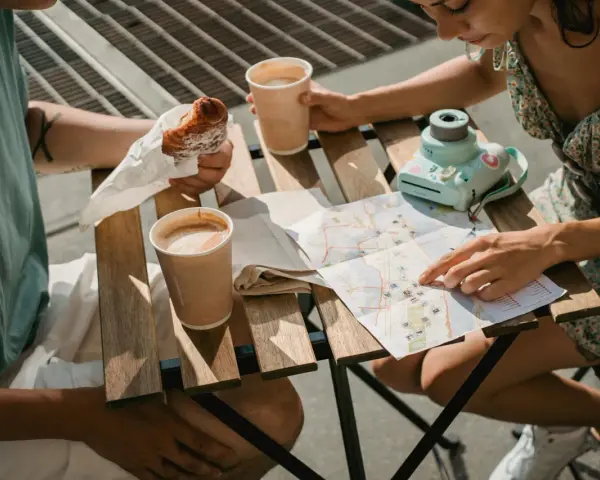 Para planuje podróż, studiując mapę i jedząc croissanty. Na stole kawa i aparat.
