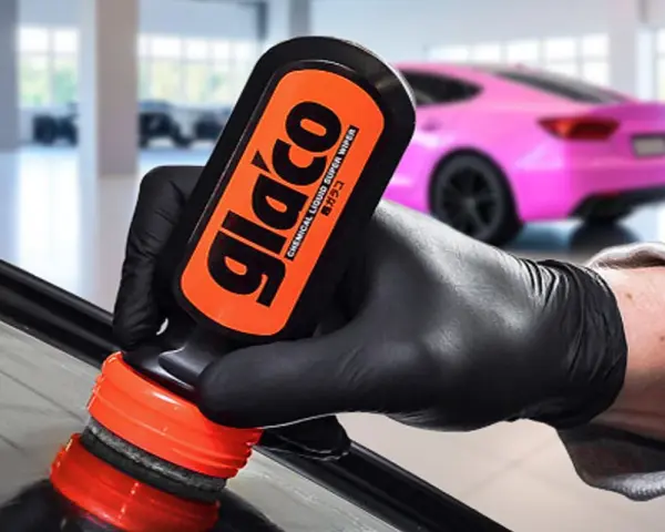 En hand med svart handske applicerar glasförsegling från en Glaco-flaska på en bilruta. En rosa bil syns i bakgrunden.
