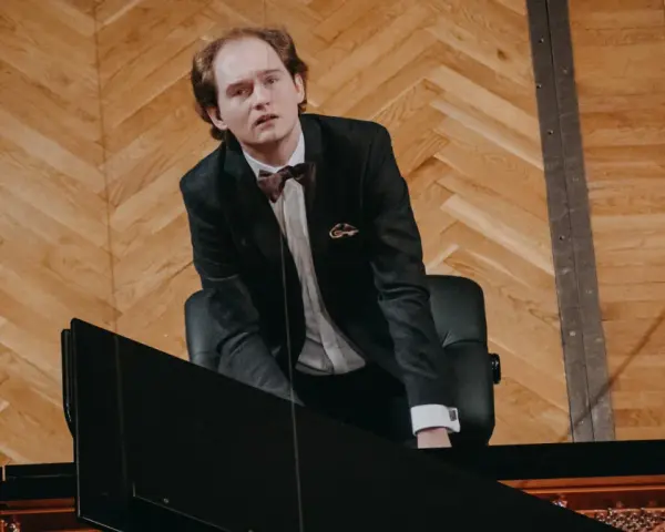 Piotr Ryszard Pawlak w eleganckim garniturze pochyla się nad fortepianem, gotowy do wykonania utworu.
