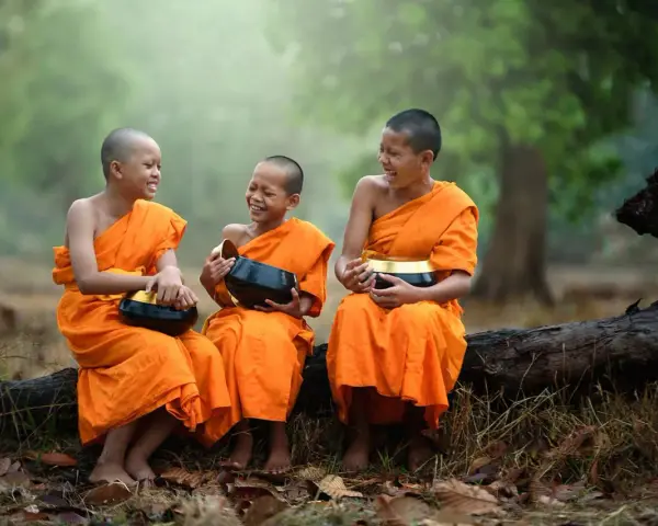 Three young monks in orange robes laugh heartily while holding alms bowls. They are sitting on a fallen log in a serene, misty forest.