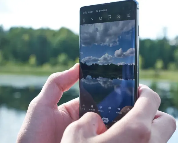 Dłonie trzymają smartfon Samsung S10, na ekranie którego widać odbicie nieba i lasu w spokojnej wodzie.