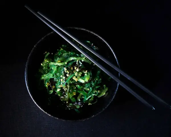 Salade de wakame algue, garnie de graines de sésame, dans un bol noir avec des baguettes.