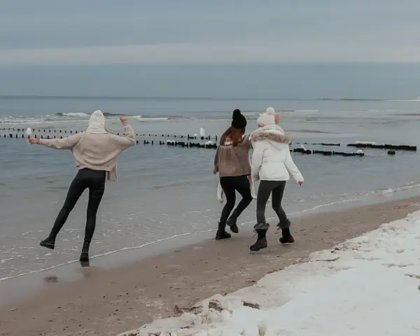 Trzy dziewczyny cieszą się zimowym dniem, gdzie zimą nad morze. Spacerują po plaży, na której leży śnieg, a fale delikatnie rozbijają się o brzeg.