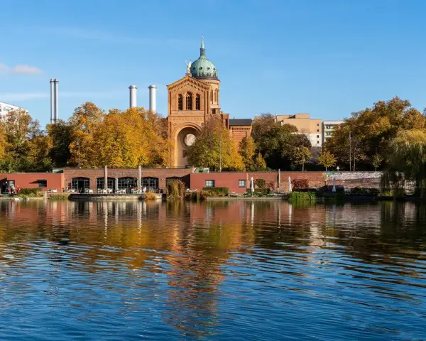 Herbstliche Bäume spiegeln sich im Wasser des Engelbeckens Berlin. Ein Café mit Außenbereich und eine Kirche mit Kuppel sind zu sehen.