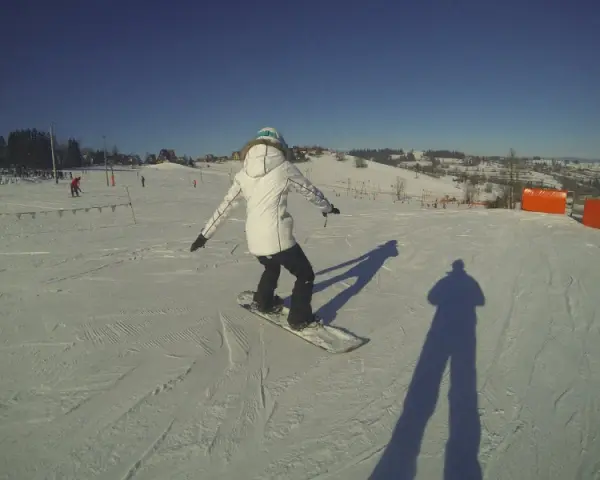 Osoba na snowboardzie na stoku narciarskim, ucząca się w szkole narciarskiej Budzyńskich. Słoneczny dzień, czyste niebo.