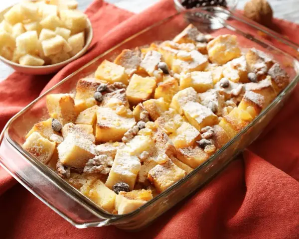 Pudding chlebowy w naczyniu, posypany cukrem pudrem i rodzynkami, obok miseczka z pokrojonym chlebem.