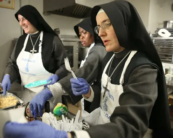 Nuns in a kitchen preparing food. One nun smiles while serving, another holds a fork, and a third looks on. This scene might answer the question: do nuns eat meat?
