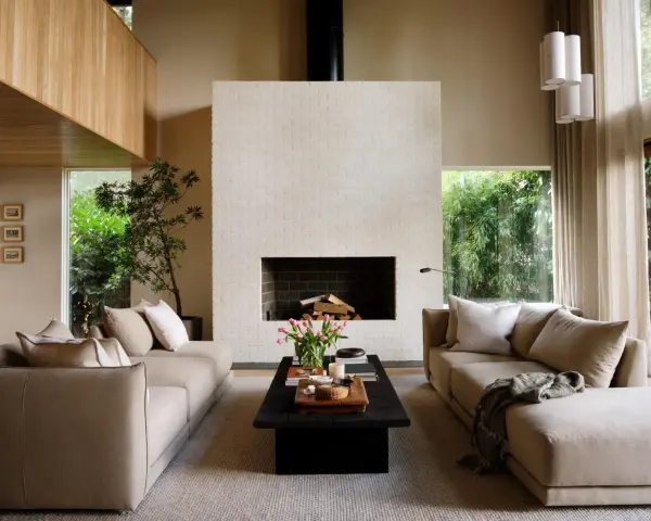 Salón de estilo japandi con sofás beige, chimenea de ladrillo blanco y mucha luz natural.