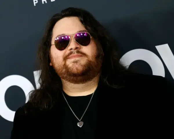 Wolfgang Van Halen w okularach przeciwsłonecznych i z naszyjnikiem w kształcie serca, na tle logo HBO.