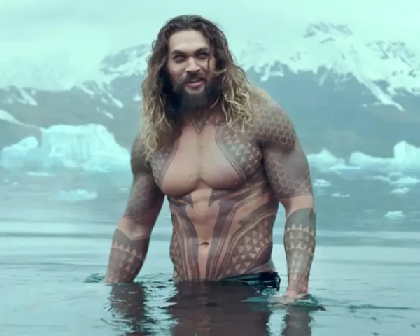 Jason Momoa w wodzie, z tatuażami, jak w filmach i serialach. W tle góry i lód.